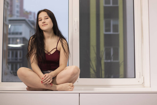 charming brunette woman sitting on windowsill. morning routine, coziness, city life. copy space  - Foto, Imagem