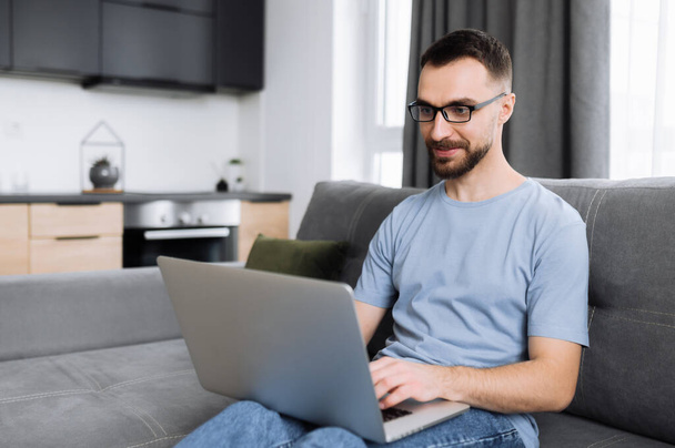 Confident male freelancer working on project from home, searching information in the network. Caucasian male manager or IT specialist sitting on the couch, using laptop, chatting with friends or - Foto, afbeelding