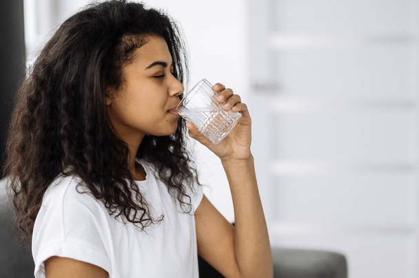 Een gezonde levensstijl. Afro-Amerikaanse jonge vrouw drinkt een glas water terwijl ze thuis op de bank zit. Mooie gezonde vrouw volgen gezonde levensstijl - Foto, afbeelding