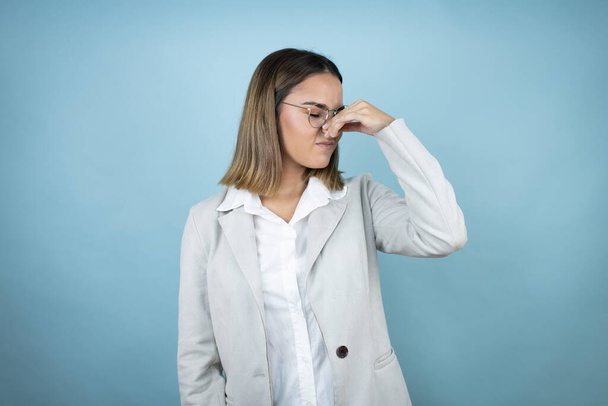 Young business woman over isolated blue background smelling something stinky and disgusting, intolerable smell, holding breath with fingers on nose - Photo, Image