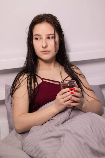 charmante slaperige brunette vrouw met glazen kopje thee in bed met grijs beddengoed. Morgen routine. kopieerruimte - Foto, afbeelding