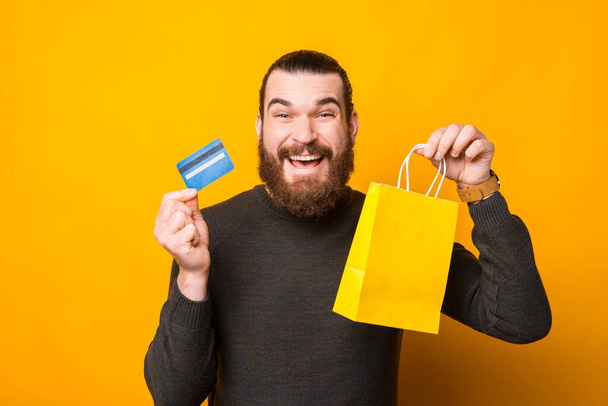 Fénykép boldog szakállas férfi kezében hitelkártya és sárga bevásárló táska, online vásárlás - Fotó, kép