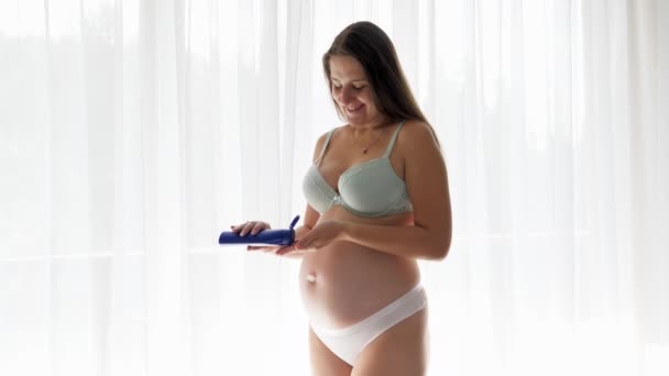 Glücklich lächelnde Schwangere, die ein Baby erwartet und Feuchtigkeitscreme oder Lotion auf ihre Körperhaut aufträgt. Konzept von Schönheit, Schwangerschaft und Gesundheitsfürsorge. - Filmmaterial, Video