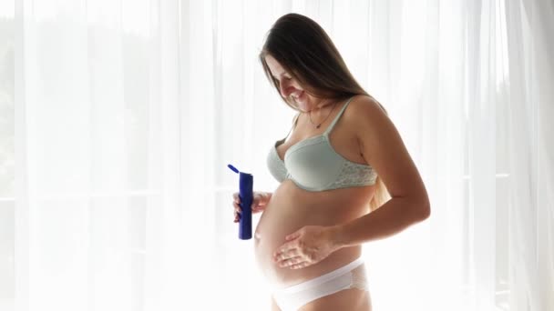 Šťastná usměvavá těhotná žena aplikující anti strie krémem z láhve. Koncept krásy, těhotenství a péče o pleť. - Záběry, video