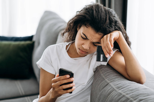 Upset girl sitting on the couch, having problems with payment by credit card. Young african american female use smartphone, make online shopping, and have problems with internet connection or online - Фото, изображение