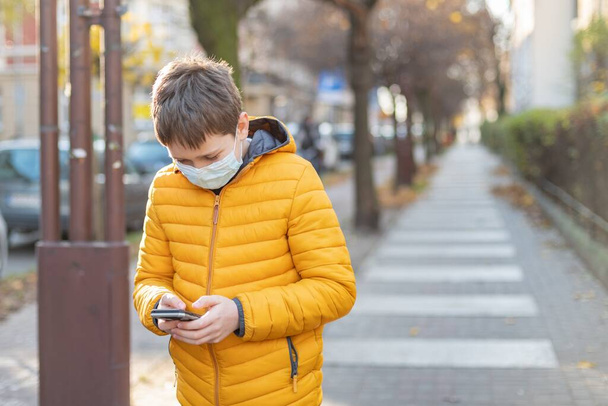 Ένα αγόρι με προστατευτική μάσκα στέκεται στο δρόμο κρατώντας ένα τηλέφωνο στο χέρι του. - Φωτογραφία, εικόνα