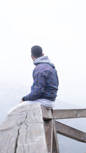 De jonge wandelaar zit op een houten hek naast een met mist bedekte berg. - Foto, afbeelding
