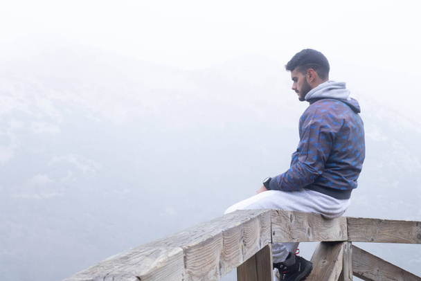 Der junge Wanderer sitzt auf einem Holzzaun neben einem nebelbedeckten Berg. - Foto, Bild