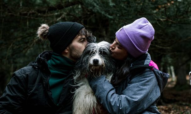 Nice portrait of a young couple of walkers kissing on the cheek of their shepherd dog. They are in a forest next to the mountain. - Photo, Image