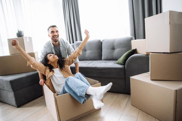 Счастливая многорасовая пара веселится с картонными коробками в своем новом доме в день переезда. Афро-американка и белый парень среди коробок в его новом доме, парень толкает девушку, пока она сидит - Фото, изображение