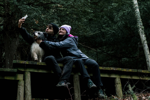 The young couple are sitting down and take out a selfie on a wooden plank road in the forest. Next to them is their shepherd dog. - Photo, Image