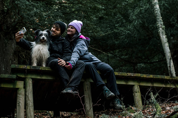 Улыбающаяся пара садится и делает селфи на деревянной дороге в лесу. Рядом с ними их пастушья собака.. - Фото, изображение