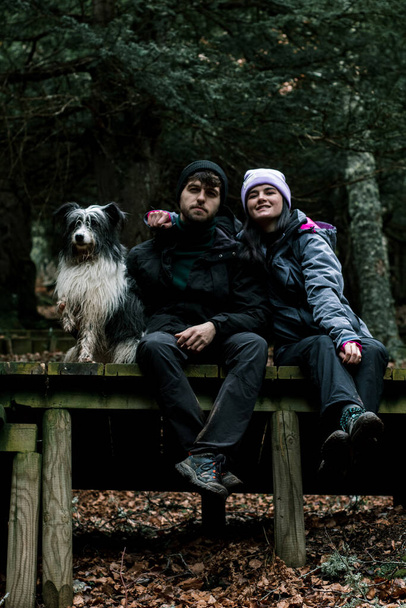 Das junge Paar sitzt auf einer Holzplanke im Wald. Neben ihnen steht ihr Schäferhund.. - Foto, Bild