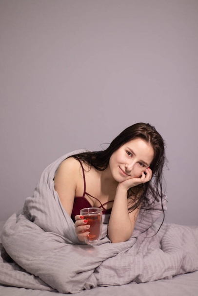 Yatağında cam bardak çay ve gri çarşaflı çekici, esmer kadın. Sabah rutini. Boşluğu kopyala - Fotoğraf, Görsel