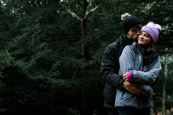 Хороший снимок молодой белой пары, целующей друг друга в щеку в лесу. Они одеты в зимнюю одежду. - Фото, изображение