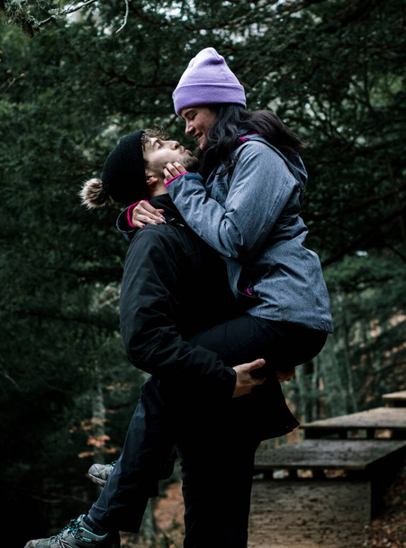 Die junge Kaukasierin mit dem rosafarbenen Hut küsst ihren Freund, während er sie in seinen Armen hält. Sie befinden sich auf einem schönen Holzweg an einem Bergpfad. - Foto, Bild