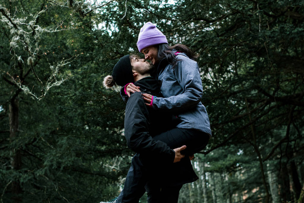 The young Caucasian woman in the pink hat kisses her boyfriend while he holds her in his arms. They are on a beautiful wooden path by a mountain trail. - Photo, Image