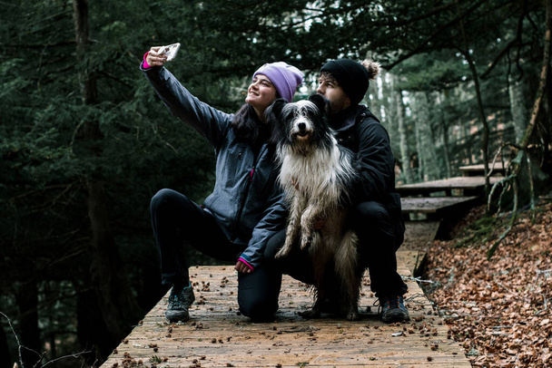 The young couple of hikers take out a selfie with their shepherd dog on a nice wooden path next to the forest - Photo, Image