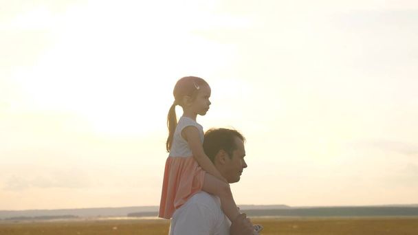 Папа с дочерью на плечах ходит на солнце. Ребенок гуляет с папой на закате. Счастливая семья отдыхает в парке. Отец носит ребенка на плечах, идя на свежем воздухе. Семейный выходной - Фото, изображение