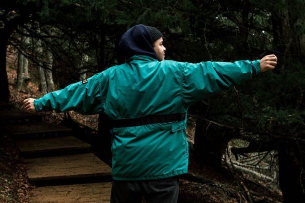 El joven excursionista con abrigo azul y capucha extiende sus brazos mientras camina por un sendero de madera entre los árboles - Foto, imagen