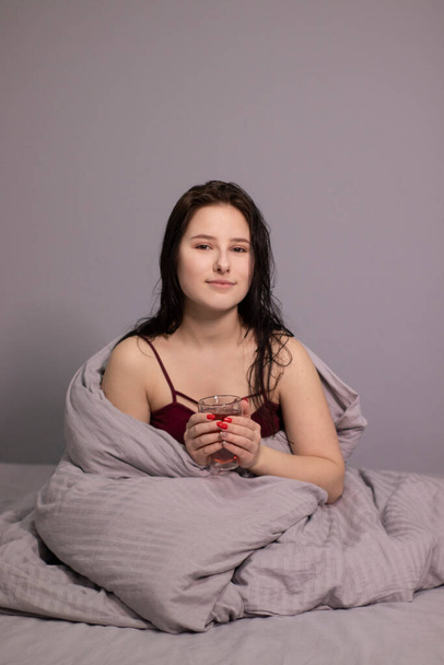 charmante slaperige brunette vrouw met glazen kopje thee in bed met grijs beddengoed. Morgen routine. kopieerruimte - Foto, afbeelding