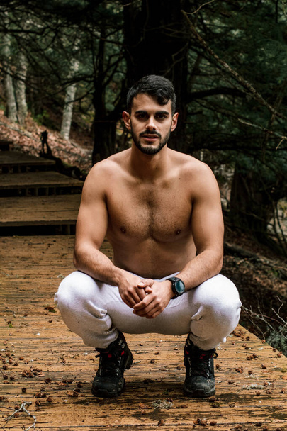 Der junge muskulöse Wanderer posiert in der Hocke. Es liegt auf einem Holzweg neben dem Berg. - Foto, Bild