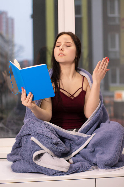 elbűvölő barna nő kék takaróba csomagolva ül az ablakpárkányon kék noteszben. Reggeli rutin, meghittség, olvasás, városi élet. másolás helye  - Fotó, kép