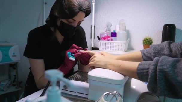 Master of manicure in protective cap and nitrile gloves sits in salon and under table lamp makes hardware manicure. Miller cleans nails of client who holds his hands on  armrest above vacuum cleaner - Footage, Video