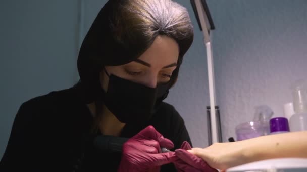 The master of manicure dressed according to all the rules in a mask, gloves and hat in a beauty salon sits at a table and under a table lamp with nail file carefully makes neat shape of client's nails - Video