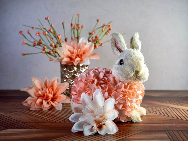Conejo de Pascua cubierto de pétalos de flores de melocotón con sus orejas en el aire sentado en un mostrador de madera con un jarrón de flores de melocotón en el fondo y más flores en primer plano. - Foto, Imagen