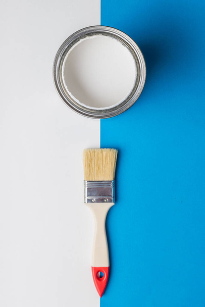 Ένα πινέλο και ένα ανοιχτό κουτί λευκής μπογιάς στο περίγραμμα των λευκών και μπλε χρωμάτων. Εκτέλεση έργων ζωγραφικής. - Φωτογραφία, εικόνα
