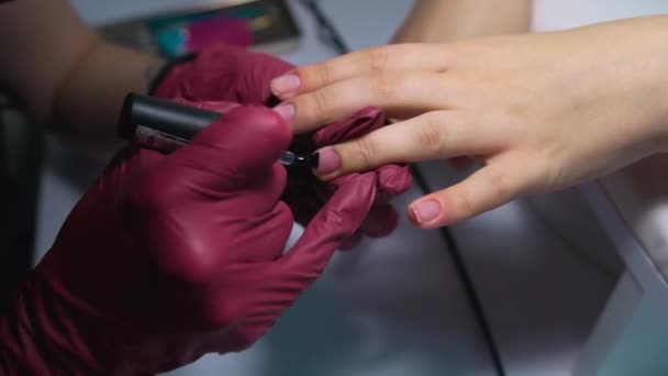 Manicurist in a special form and in disposable nitrile gloves with a black brush applies a transparent base under the gel on the nails of the client in a beauty salon - Footage, Video
