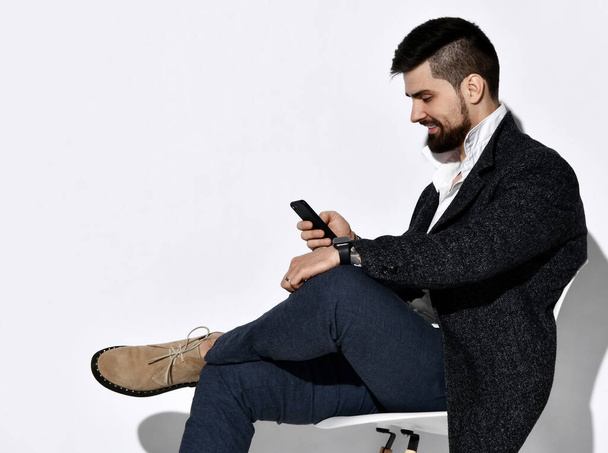  Uśmiechnięty brodaty mężczyzna w dżinsach i stylowa, luźna kurtka tweedowa używa wiadomości ze smartfona. Widok boczny - Zdjęcie, obraz