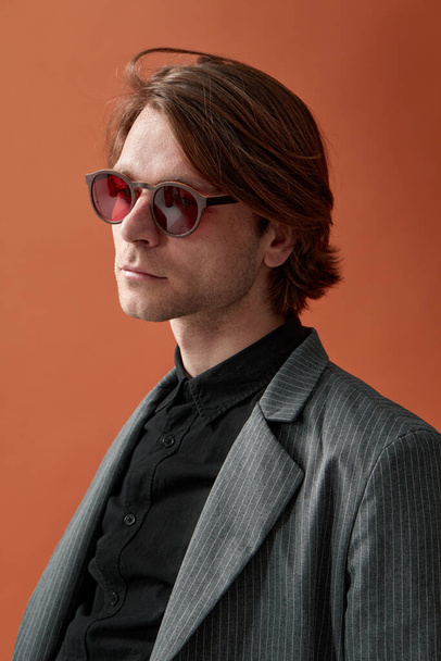 Aantrekkelijk elegant mannelijk model in rode bril, staande met een stevige houding op bruine studioachtergrond. - Foto, afbeelding