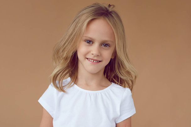 Retrato frontal de niña bonita con cabello rubio ondulado, vestida con ropa blanca, sobre fondo beige. - Foto, imagen