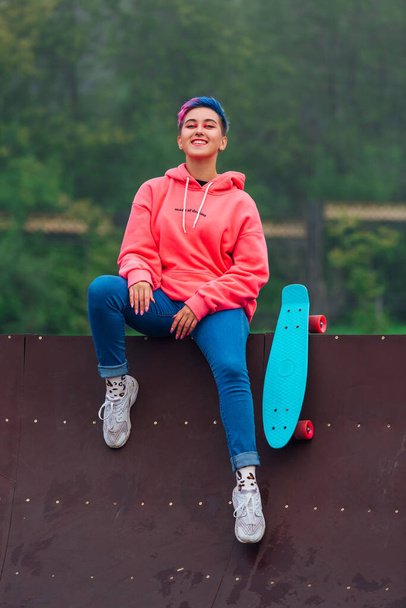 Portret van een trendy mooi jong meisje met kort gekleurd haar en neus piercing gekleed in roze hoodie en jeans zitten naast de skateboard court met haar blauwe plastic skateboard. - Foto, afbeelding