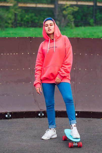 Portrait of a trendy pretty young girl dressed in pink hoodie and jeans standing next to the skateboard court with her blue plastic skateboard. - Foto, immagini