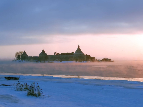 χιονισμένη ακτή και Oreshek Φρούριο στην ομίχλη στο νερό της λίμνης στο φως της ανατολής - Φωτογραφία, εικόνα