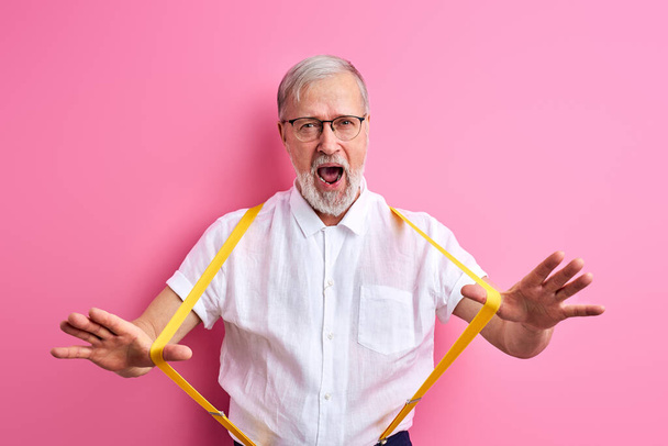 handsome senior man holding suspenders isolated on pink background - Фото, изображение