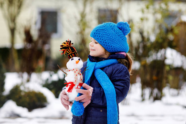 Мила маленька дівчинка робить міні сніговика і їсть морквяний ніс. Чарівна здорова щаслива дитина грає і розважається зі снігом, на відкритому повітрі в холодний день. Активний відпочинок з дітьми взимку
 - Фото, зображення
