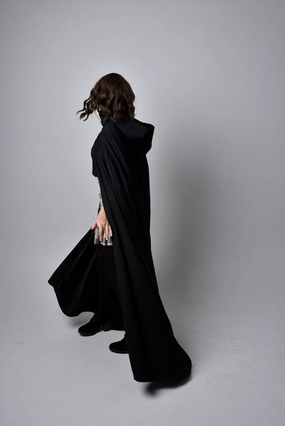 Ganzkörperporträt einer hübschen brünetten Frau, die Schottenrock und Stiefel mit langem schwarzen Mantel trägt. Stehende Pose mit einem Zauberstab vor Studiohintergrund. - Foto, Bild