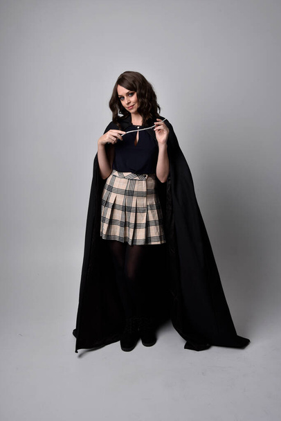 full length portrait of pretty brunette woman wearing tartan skirt and boots with long black cloak.. Standing pose holding a magical wand against a  studio background. - Foto, afbeelding