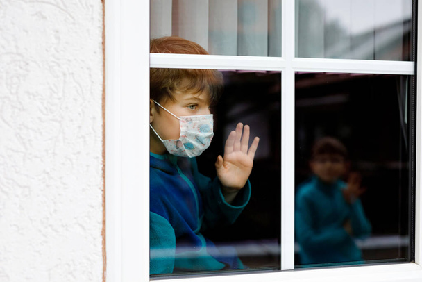 Lovely little school kid boy by a window wearing medical mask at home or school during pandemic coronavirus quarantine. Lonely upset child looking at best friend and wafes. - Fotó, kép