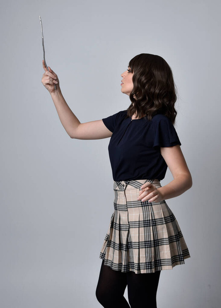 full length portrait of pretty brunette woman wearing tartan skirt and boots with long black cloak.. Standing pose holding a magical wand and casting a spell against a  studio background. - Valokuva, kuva