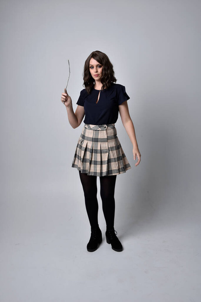 full length portrait of pretty brunette woman wearing tartan skirt and boots with long black cloak.. Standing pose holding a magical wand and casting a spell against a  studio background. - Photo, image