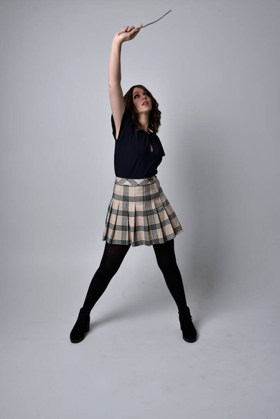 full length portrait of pretty brunette woman wearing tartan skirt and boots with long black cloak.. Standing pose holding a magical wand and casting a spell against a  studio background. - Foto, Imagen