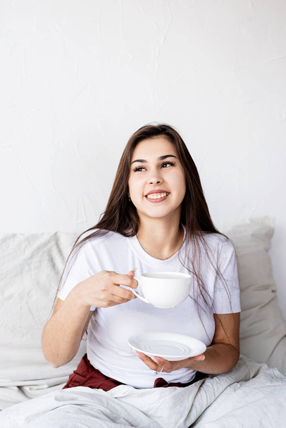 Valentines Day. Young brunette woman sitting awake in the bed with red heart shaped balloons and decorations drinking coffee eating croissants - Фото, изображение