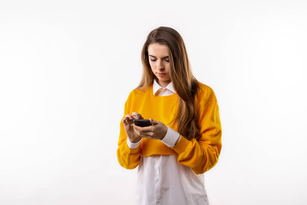 Uśmiechnięta młoda brunetka w luźnych ciuchach używa telefonu komórkowego, wpisując SMS-y. Technologia, młodzież i koncepcja komunikacji. Studio shot, białe tło - Zdjęcie, obraz