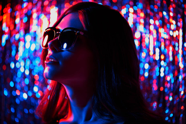 Fashion portrait of young elegant trendy woman in sunglasses. Neon light. Colored glitter tinsel background - Foto, immagini