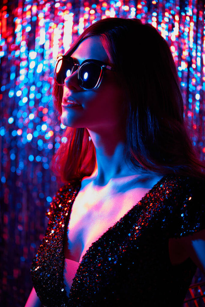 Fashion portrait of young elegant trendy woman in sunglasses. Neon light. Colored glitter tinsel background - Foto, Imagen